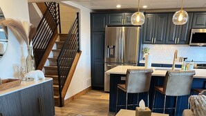Una sala moderna con un sofá de cuero, un comedor con mesa y sillas, una cocina con armarios oscuros y electrodomésticos de acero inoxidable, y una escalera con barandilla negra.