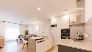 Een moderne keuken met een inbouw vaatwasser, een magnetron en een fornuis. Er is een eethoek met een tafel en stoelen, en een woonkamer met een bank en een plant.