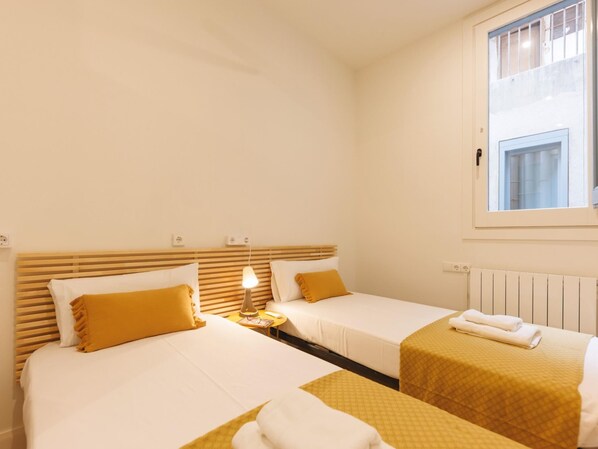 A hotel room with two beds, each with a wooden headboard, white bedding, and a yellow accent pillow. There is a bedside lamp on one bed, and a window with blinds on the wall. - Bravissimo Ciutadans Penthouse, with 3 bedrooms (Girona)