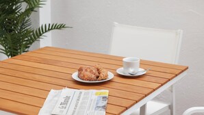 Une table en bois sur laquelle se trouvent une assiette de croissants et une tasse de café, un journal et une chaise blanche.