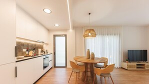 Une cuisine moderne avec des meubles blancs, une table à manger en bois avec quatre chaises, un luminaire suspendu et un téléviseur dans l’espace de vie.