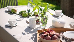 Une table d’extérieur recouverte d’une nappe blanche, avec une carafe, un vase rempli de verdure, des pommes et un plateau de prunes.