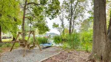 A wooden swing set, a small boat, and a gravel path in a wooded area with trees and a body of water in the background.