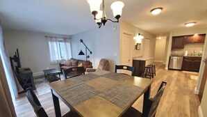 A dining area with a table and chairs, a chandelier, and a kitchen in the background.