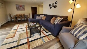 Een woonkamer met een bank, een bijzettafeltje en een kamerkleed. Achtergrond toont een eethoek met stoelen en een tafel.