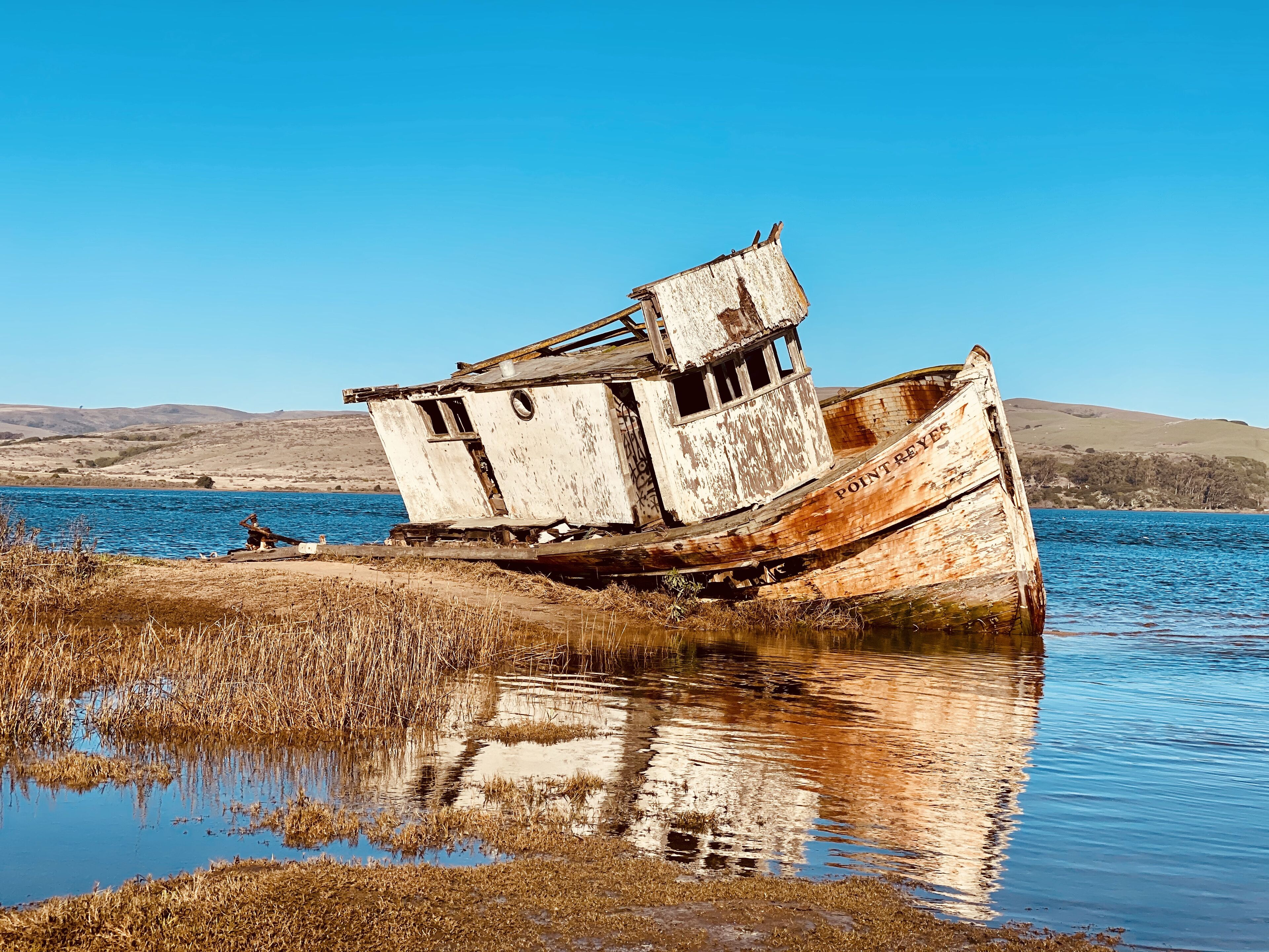 An abandoned shipwreck with a cabin structure, situated in a calm body of water with a clear sky and distant hills in the background.