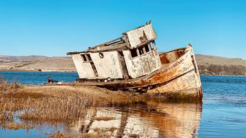 An abandoned shipwreck with a cabin structure, situated in a calm body of water with a clear sky and distant hills in the background.