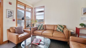 A living room with a brown leather sofa, a glass coffee table with a book, and large windows with wooden frames. - Limantour Lodge-Laguna House-Perfect for Families! (Point Reyes Station)
