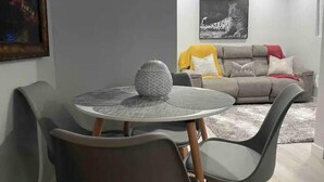 Een eethoek met een ronde tafel en stoelen, een grijze bank met kussens, en een lijst met een afbeelding aan de muur.