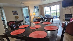 Un coin repas avec une table en bois foncé, des sets de table rouges et des chaises noires. Un téléviseur est fixé au mur et une cheminée se trouve au fond de la pièce.