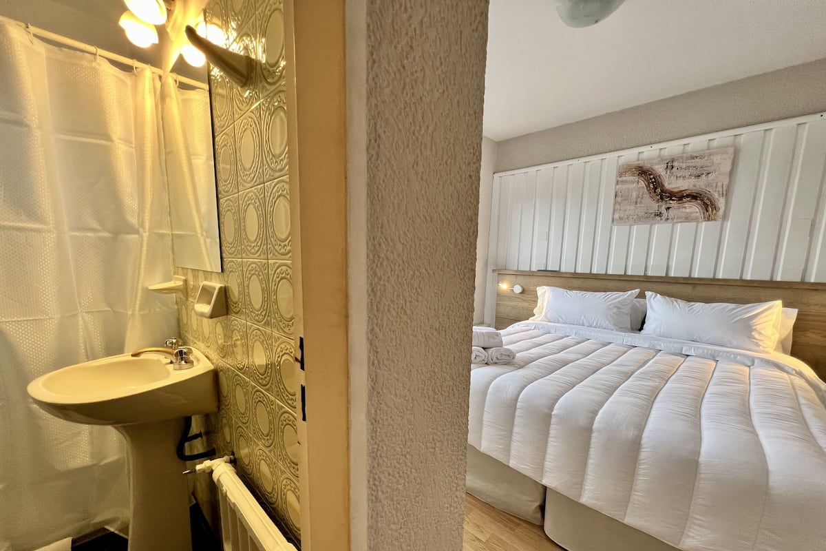 A hotel room with a bathroom, a bed with white bedding, and a wall-mounted light fixture.