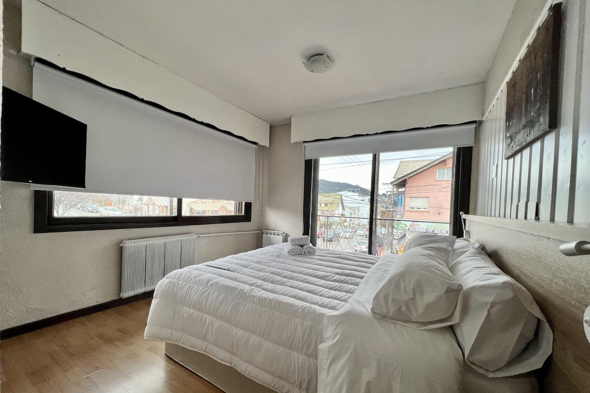 A bedroom with a large bed, a window with a view of buildings, and a ceiling fan.