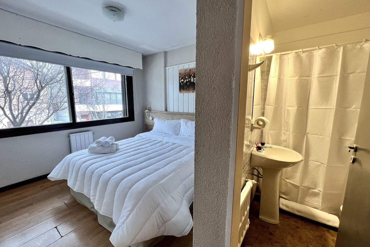 A bedroom with a large bed, a window, a radiator, and a bathroom with a sink and shower curtain.
