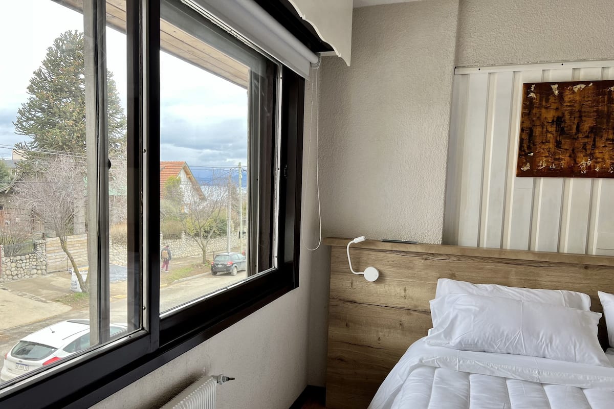 A bedroom with a large window, a radiator, a wooden headboard, and a white bed.