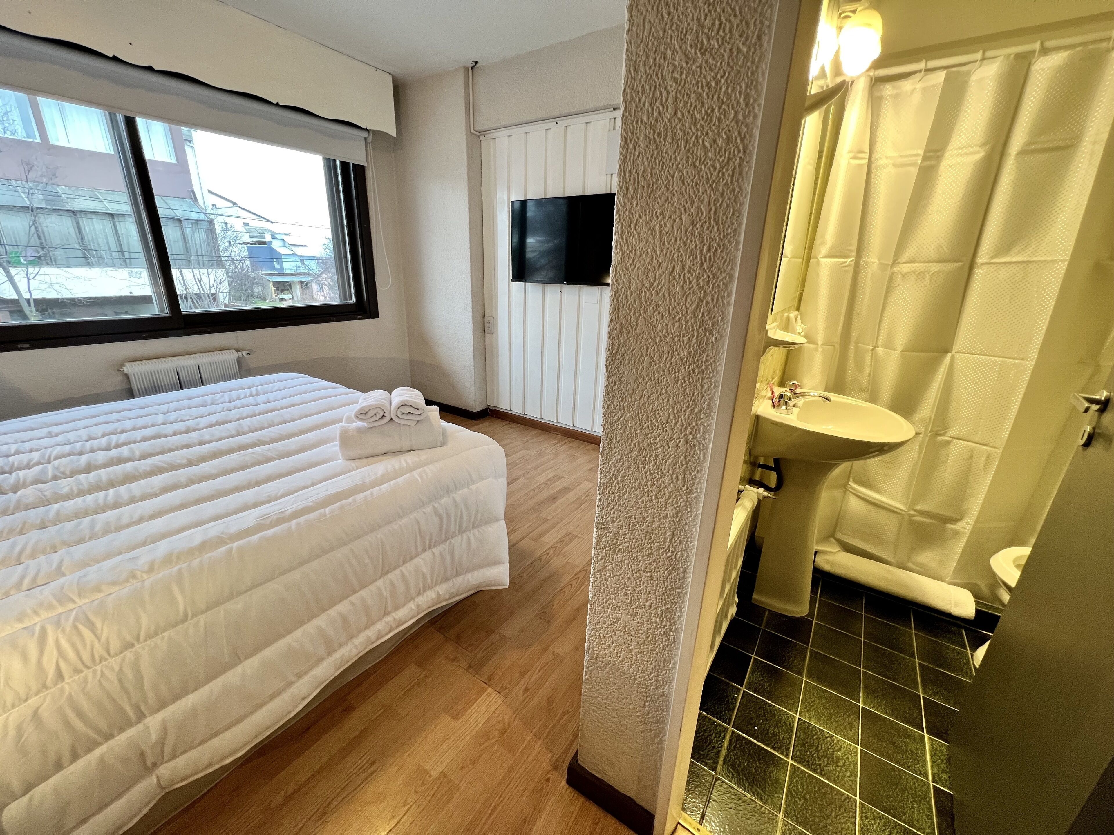 A hotel room with a large bed, a bathroom with a sink and shower, and a TV on the wall.