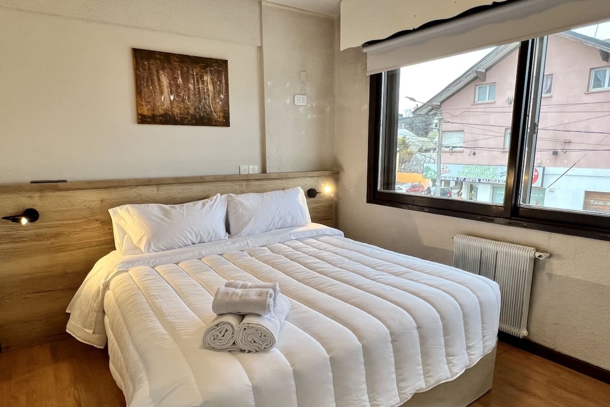 A neatly made bed with white bedding and rolled towels, a wooden headboard, a wall-mounted light, and a window with a view of buildings outside.