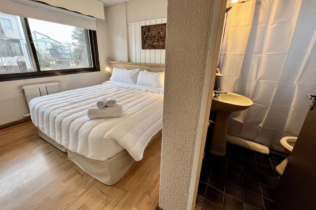 A hotel room with a bed, a window, a radiator, a bathroom with a sink and toilet, and a shower curtain.