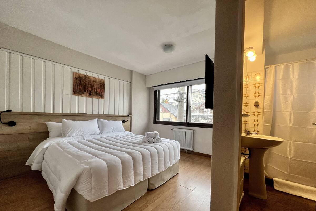 A bedroom with a large bed, a window, a radiator, and a bathroom with a sink and mirror.