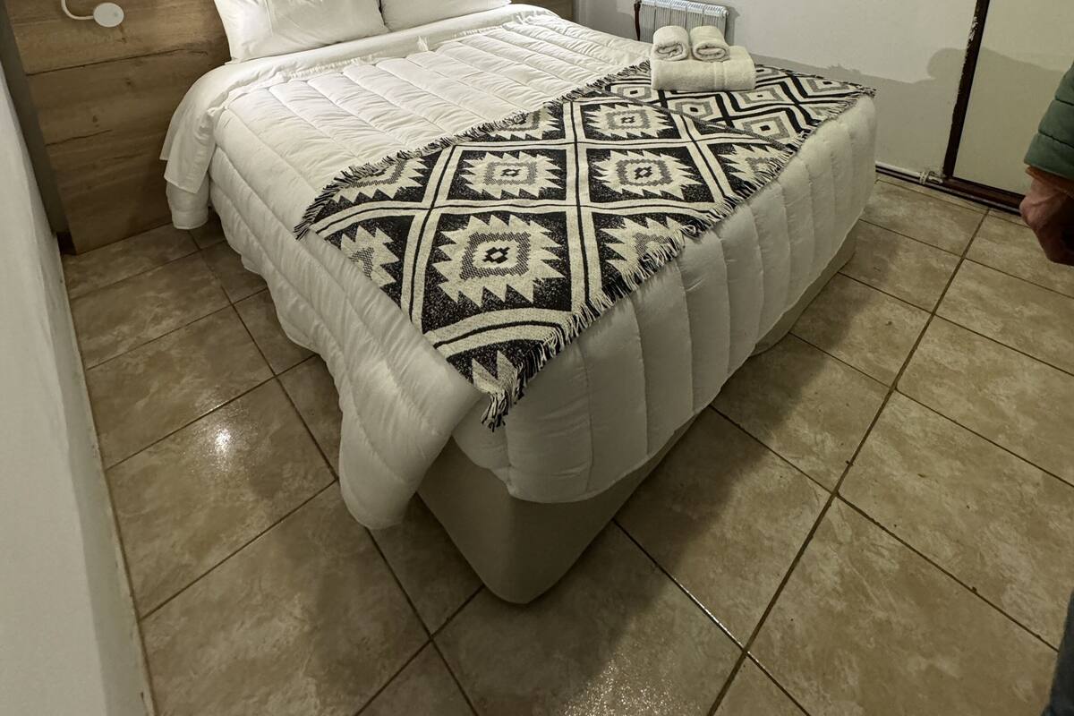A bedroom with a bed, a nightstand, a wall-mounted light, and a window with blinds.