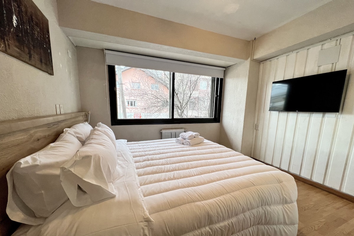 A bedroom with a large bed, a window with blinds, a radiator, and a flat-screen TV mounted on the wall.