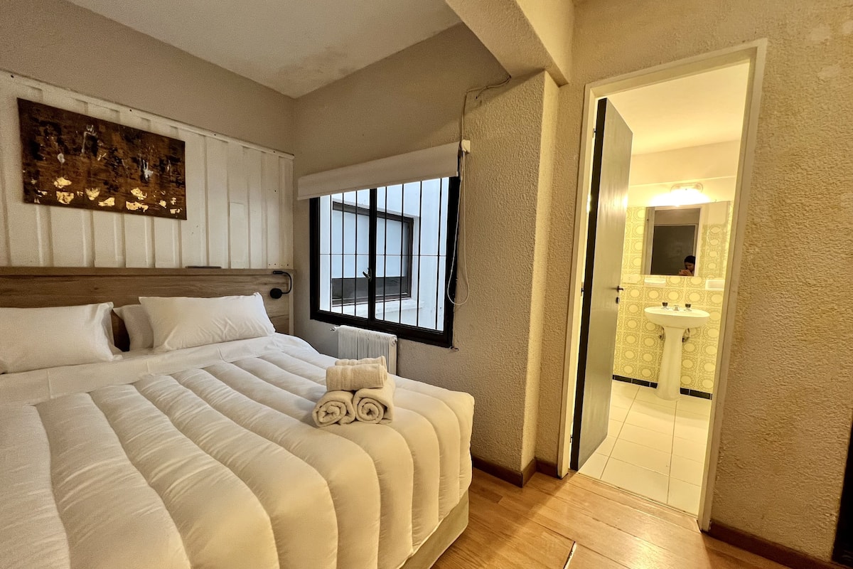 A bedroom with a large bed, a window, a radiator, and a bathroom visible through an open door.