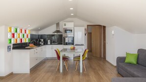 A modern kitchen with white cabinets, a dining table with colorful chairs, and a grey sofa in a spacious living area. - Vacations in Funchal, sea view - Casa Strelitzia II (Funchal)