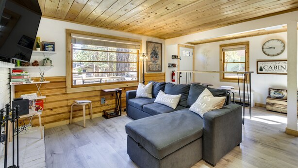 A cozy cabin interior with a sectional sofa, wooden floors, and a large window offering a view of the outdoors. - Sugarloaf Vacation Rental ~ 7 Mi to Big Bear Lake! (Sugarloaf)