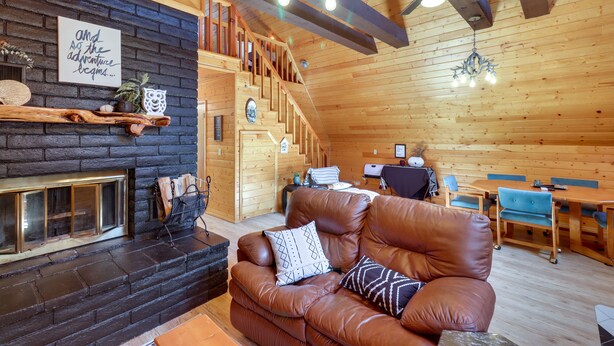 A cozy cabin interior with a fireplace, leather sofa, wooden dining table, and a staircase leading to an upper level. - Decks + Fire Pit: Rustic-Modern Overgaard Cabin! (Overgaard)
