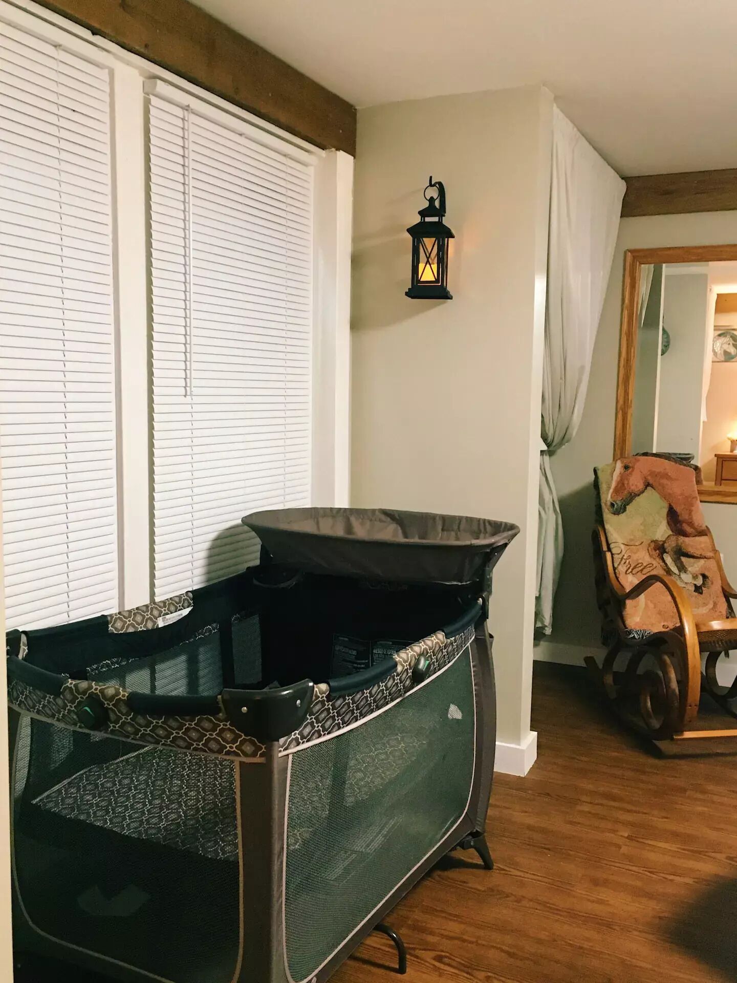 Una habitación con un ventanal con persianas, un mecedora infantil negra con estampado de jirafa, una mecedora de madera y un farol colgante en la pared.