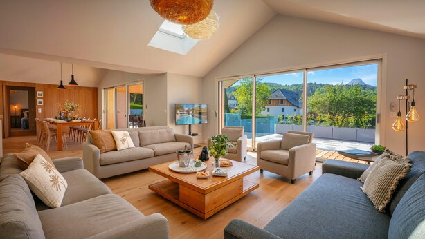 En rummelig stue med højt til loftet, et spiseområde og store skydedøre, der fører ud til en balkon med udsigt til et bjerglandskab.