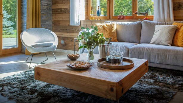 Ein gemütliches Wohnzimmer mit Couch, Sessel und einem Holztisch. Durch die Fenster ist ein Blick auf Bäume und Berge möglich.