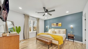 Ein Schlafzimmer mit Bett, einer Bank, einem Nachttisch, einem Deckenventilator, einem Fernseher und einem Fenster mit Vorhängen.