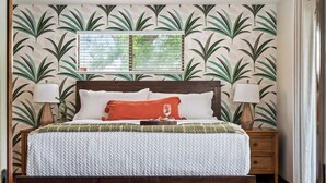 Une chambre à coucher comprenant un lit, deux tables de chevet, une fenêtre avec des stores et un mur orné d’un motif de feuilles.