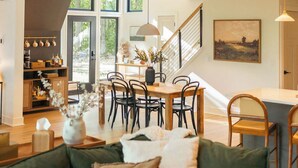 A spacious loft with a high ceiling, wooden beams, a dining area with a large table and chairs, a cozy seating area with a green sofa, and a staircase leading to an upper level. - Modern Aframe with River Views, Sauna, Hot Tub (Fennville)