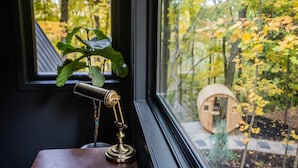 A brass desk lamp on a wooden table by a window with a view of a wooden structure and autumn foliage. - Modern Aframe with River Views, Sauna, Hot Tub (Fennville)