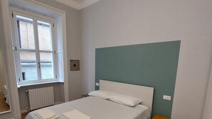 Uma cama bem arrumada com lençóis e almofadas brancas, uma mesinha de cabeceira e uma janela com persianas.