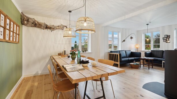 Een ruime woonkamer met een houten eettafel, een zwarte leren bank en een groot raam met uitzicht op de omgeving.