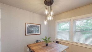 Un coin repas avec une table en bois, des chaises, un luminaire suspendu, un tableau encadré et une fenêtre avec des stores.