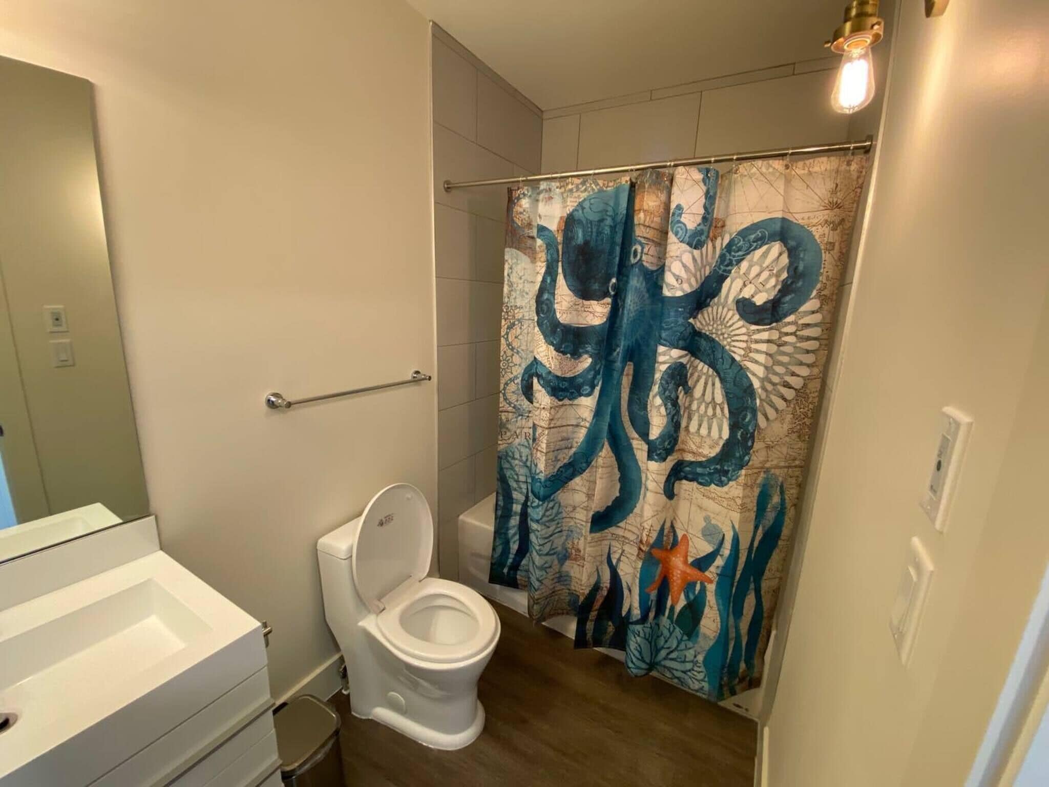 A bathroom with a white toilet, a shower curtain with a sea creature design, and a wooden floor.