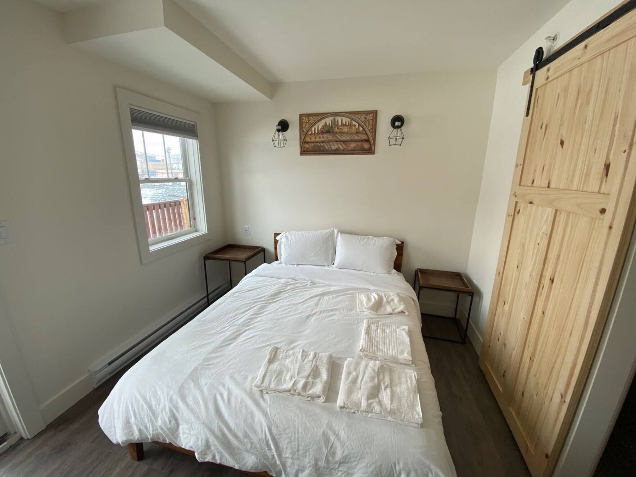 A bedroom with a large bed, two bedside tables, a window, a wooden wardrobe, and two wall-mounted lamps.