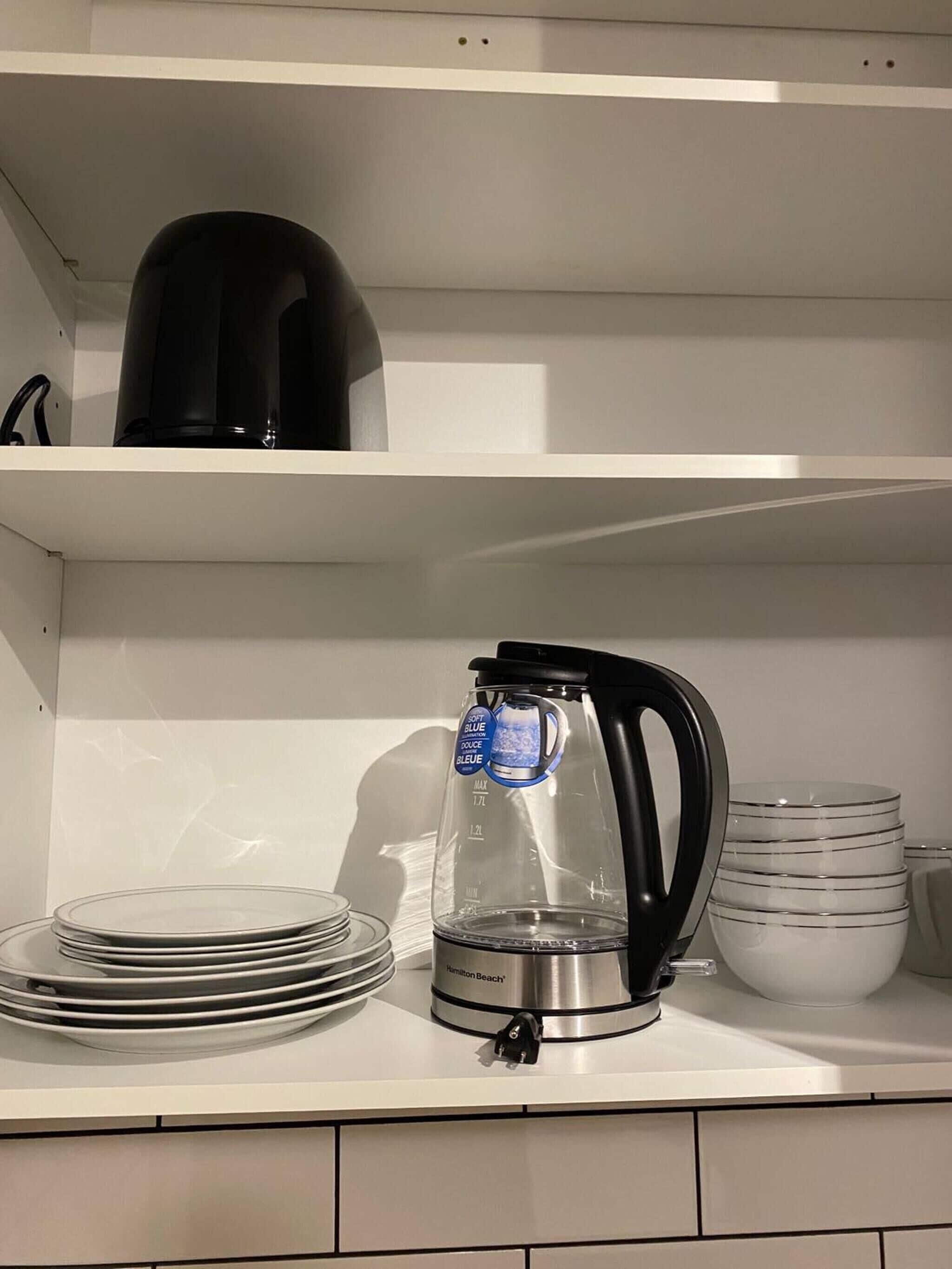 A kitchen shelf with a Hamilton Beach coffee maker, a black electric kettle, and stacked glass plates.