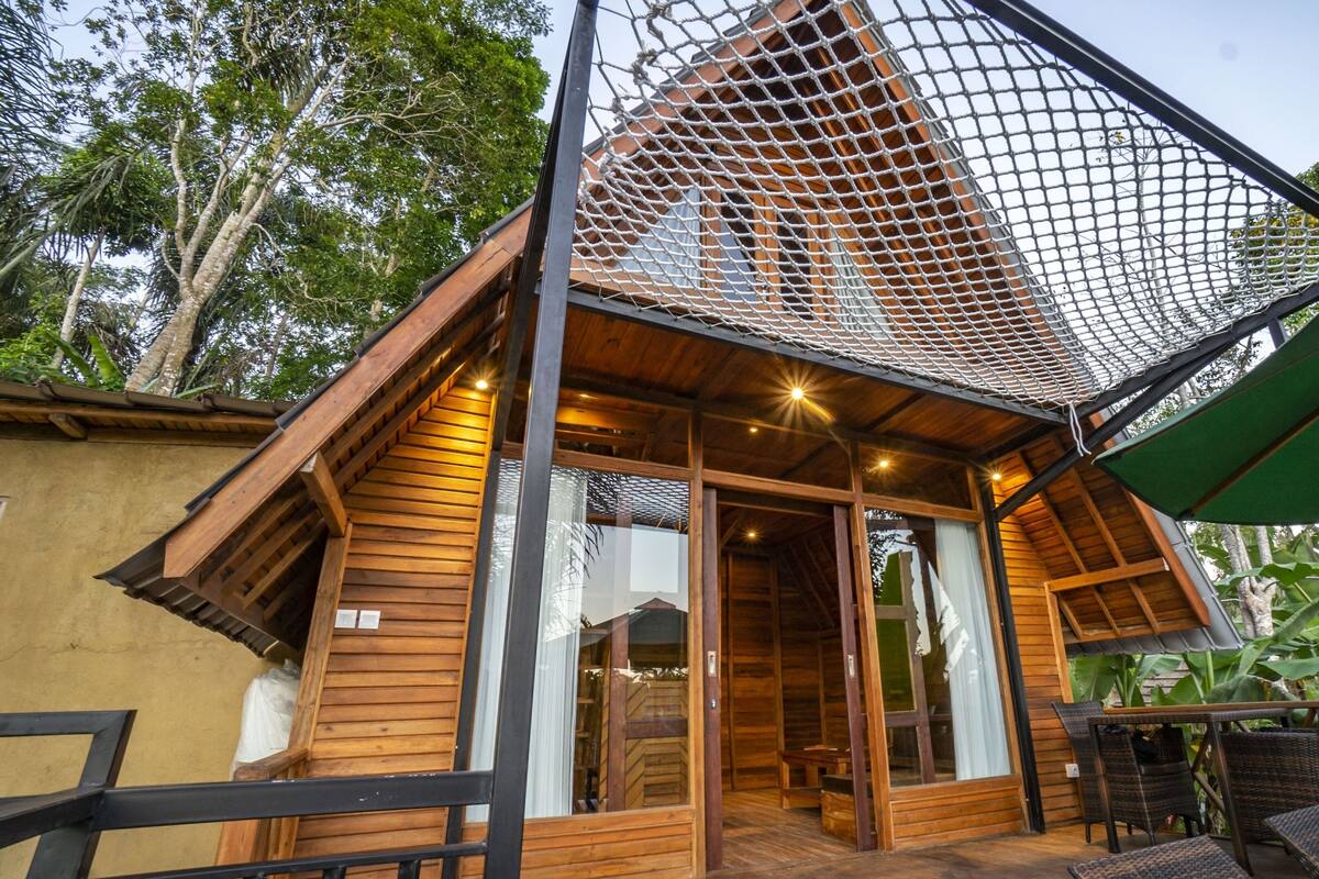 A wooden cabin with a netted roof, large windows, and a patio area with chairs and a table.