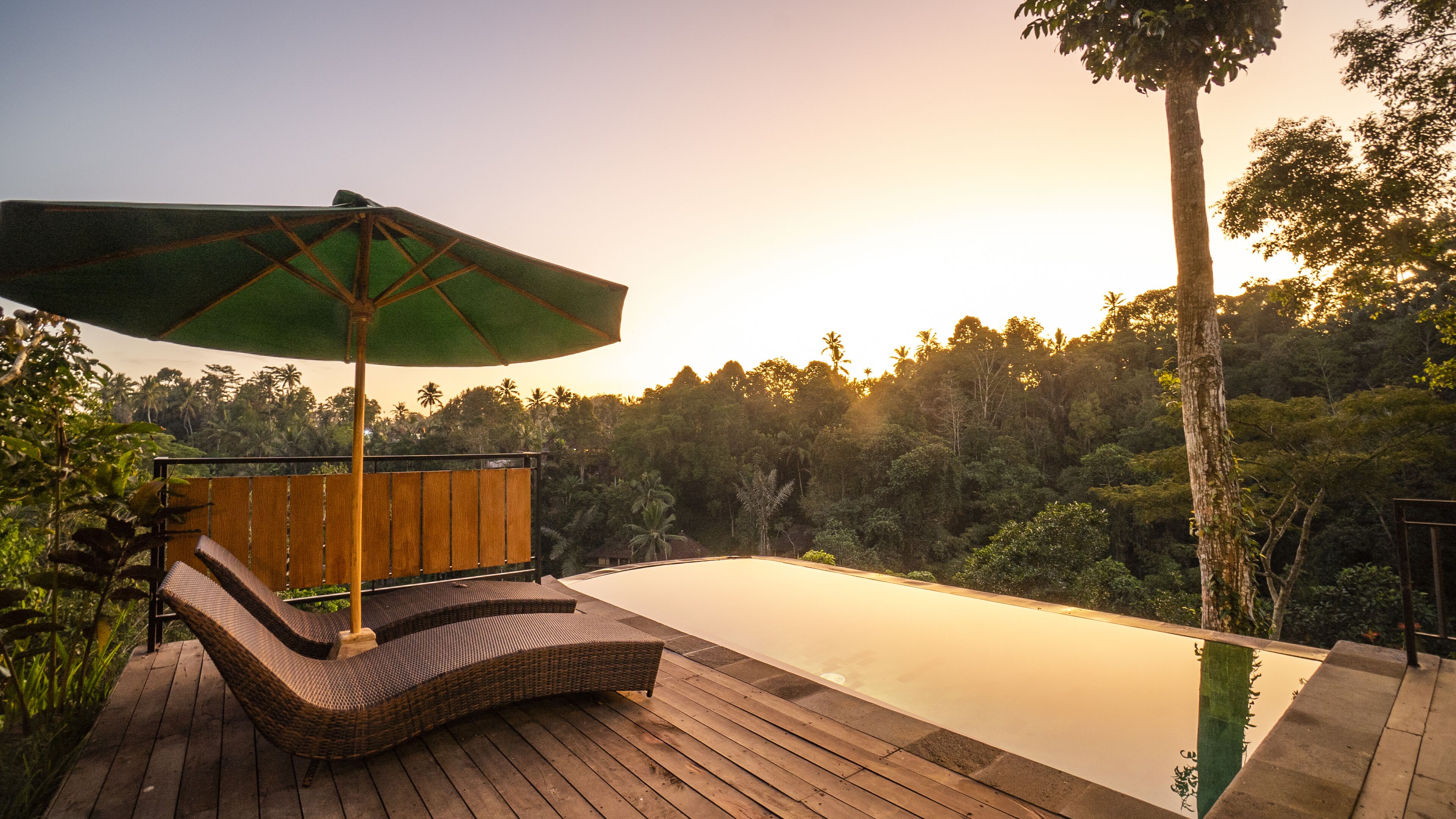 A wooden deck with a lounge chair and an umbrella, overlooking a calm infinity pool and a lush forest backdrop.
