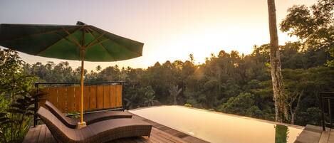 A wooden deck with a lounge chair and an umbrella, overlooking a calm infinity pool and a lush forest backdrop.