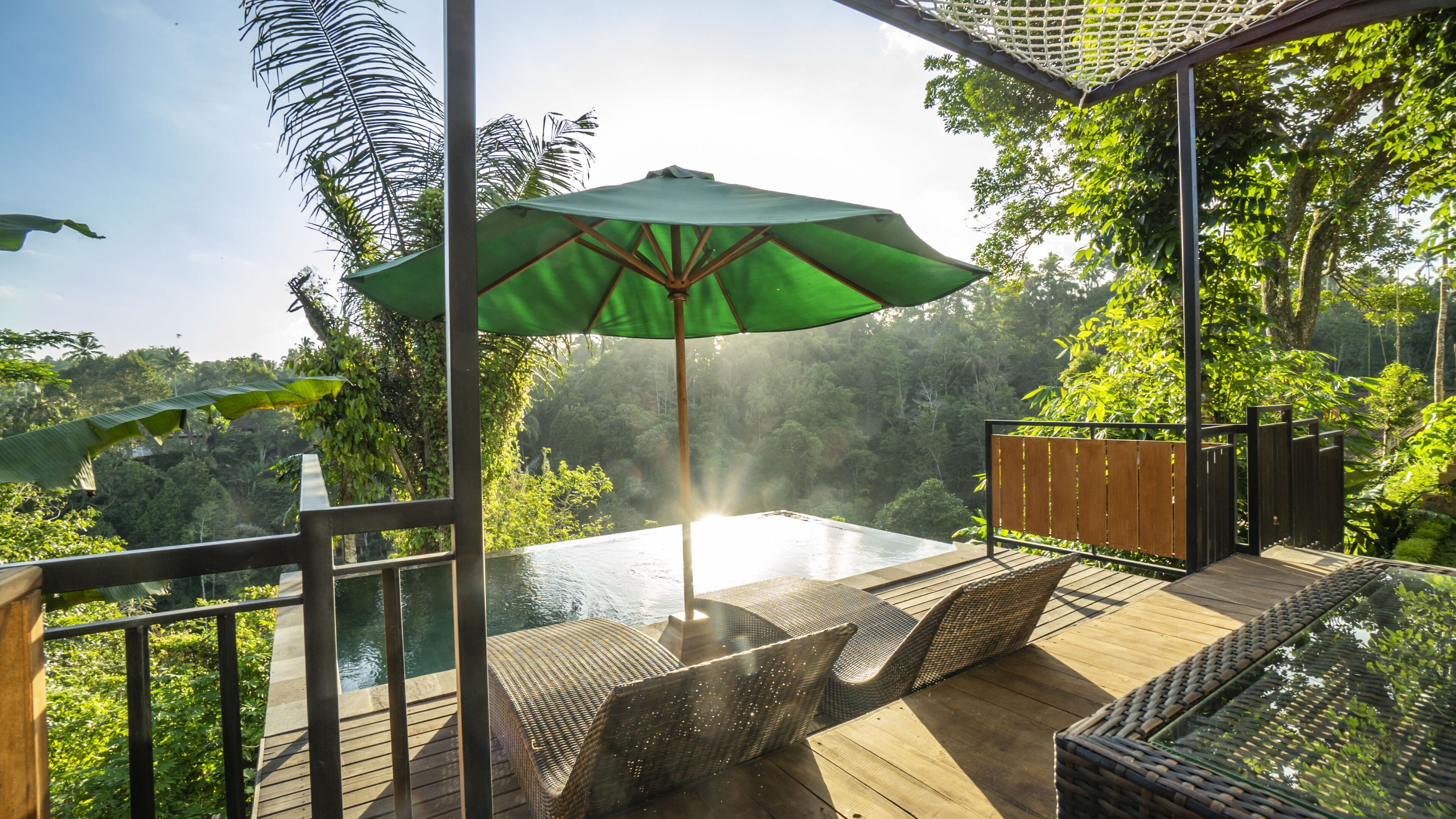 Terraza de madera con piscina de fondo de cristal, rodeada de exuberante follaje y vistas a una zona boscosa.