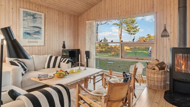 Ein gemütliches Ferienhaus-Interieur mit Holzvertäfelung, einer Couch, einem Esstisch, Stühlen, einem Kamin und einem großen Fenster mit Blick auf Bäume und ein Auto.