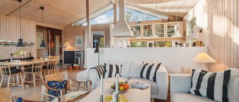 Un salon chaleureux et bien éclairé, avec un plafond en bois, un coin repas comprenant une table et des chaises, et une cuisine équipée de placards et dotée d’une fenêtre.