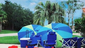 Ein Strandaufbau mit blaugrünem Sonnenschirm, Liegestühlen, einer Kühlbox, einem Spielzeugauto und einer blauen Matte.
