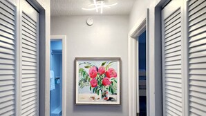 Ein Flur mit einem gerahmten Gemälde von Blumen an der Wand, einer Deckenleuchte und weißen Fensterläden an den Türen.