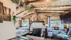 Ein gemütliches Blockhaus mit Kamin aus Stein, einem Bett mit floralem Bettbezug, einem Schreibtisch mit Computer und einer hängenden Pflanze.